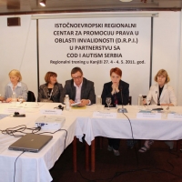 Organizational representatives are seated behind a long table with a white tablecloth. Zoltan Mihok stands to the right, speaking into a microphone.