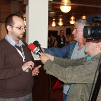 Mr. Zoltan Mihok speaks to two reporters holding microphones. A video camera at the right is filming the interview.