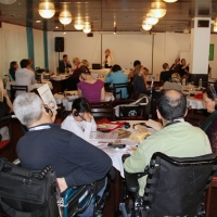 Participants are seated around tables with their backs to the camera. Marcia Rioux is speaking at the front of the room.