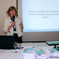 Marcia Rioux stands and speaks into a microphone during a presentation. A powerpoint presentation is projected onto the wall behind Marcia. Training materials are piled on the table in front of her.