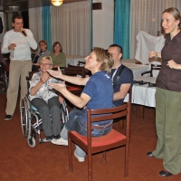 Three participants are seated and smile while a man stands and speaks into a microphone behind them. A sign language interpreter is signing in the foreground.