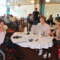 Three women are working at a table covered in water bottles, training material and electronic equipment.