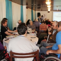 Several men and women are seated around two round tables and while Paula Pinto and Marica Rioux stand at the front of the room. A powerpoint presentation is projected onto the wall behind Paula and Marcia.