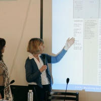 Paula Pinto and Marcia Rioux stand to the side of a slide presentation projected on the wall. Marcia Rioux is gesturing at the slide while speaking into a microphone.