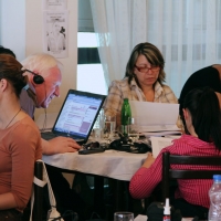 A man and three women are seated around a small round table. The man is works at a laptop computer and the women are looking at papers.