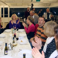 Participants sit around the dinner table in the restaurant and clap their hands while traditional musicians play instruments in the background.