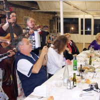 Four men play musical instruments on one side of a long table while participants smile and watch on the other side.