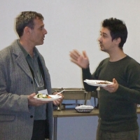 Two participants have a conversation over a bite to eat. Lunch is served in the background as the two participants stand to have their discussion. 