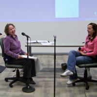Melanie and Iphigenia are seated facing each other with a microphone in front of Melanie. They are both smiling and laughing.