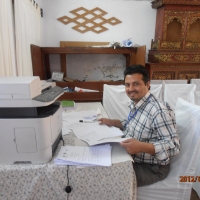 Kumar  Regmi sitting at a desk smiling at the camera working on participants lists for the country training in Nepal.  The photo is taken from his left side and he is turning his head to look at the camera.