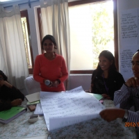 Four participants present their discussion results to the rest of the training group. Three are seated around a table and one is standing as the representative for the group. 