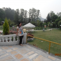 Chona stands on a patio section in front of the garden outside of the hotel. In the background there are covered seating areas, trees and grass with a path running through the middle.