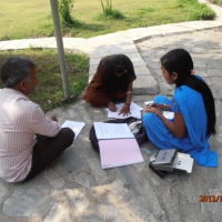 Three participants sit on the ground as the one in the centre of the photo writes and the other two (left and right in the photo) look on. 