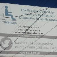 Large white sign with blue lettering saying: National Council for Persons with Physical Disabilities in South Africa. Also on the sign are the phone number, fax and email of the organization. 