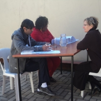<p>
	Helene Combrinck is sitting at a table being interviewed face to face by Imtiaz Moola and Zama Ngwenya.</p>
