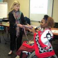 Prof. Marcia is standing and receiving a gift from Bongiwe Malope, inside the training venue. In the background there is a flip chart, tables and chairs. 