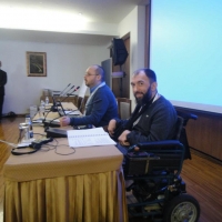 Zoltan and Samir are seated behind a table at the front of the training room , there is a large presentation screen behind them. 