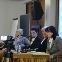 Presenters sit behind a table in the training room. They speak into microphones in front of them. A camera is visible in front of them. 