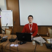 Rados sits down at the front of the room and smiles for the camera. Behind him is the screen and in front of him are the microphones and an open computer.