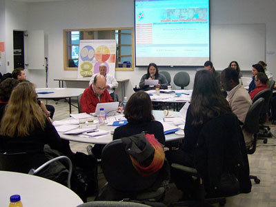 Susan Ralph is speaking into a microphone while looking at a sheet of paper in front of her. A.G.M. participatns are seated around tables in the foreground