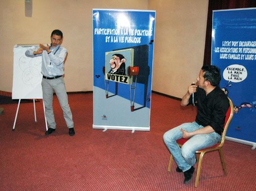 A man is seated on a chair in front of two posters dealing with participation in political life. He is speaking into a microphone and looking at a second man who is standing and using sign language