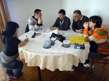 Five people are at a circular table. A woman (site coordinator) is on the left and is writing. In the middle, there are two men (monitors), one of them holding an audio recorder. The monitors are talking to a man on the right (practice interviewee). Beside this man, is a woman (personal assistant)