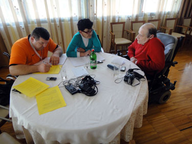 Three people are at a circular table. At the left is a man (monitor) who is taking notes. In the middle is a woman (monitor) who is speaking to a man on the right (practice interviewee).