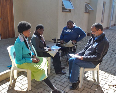 Four participants – two men and two women - are sitting in a group outside. The two women are sitting beside each other, speaking with a man who is sitting across from them. A second man is observing.